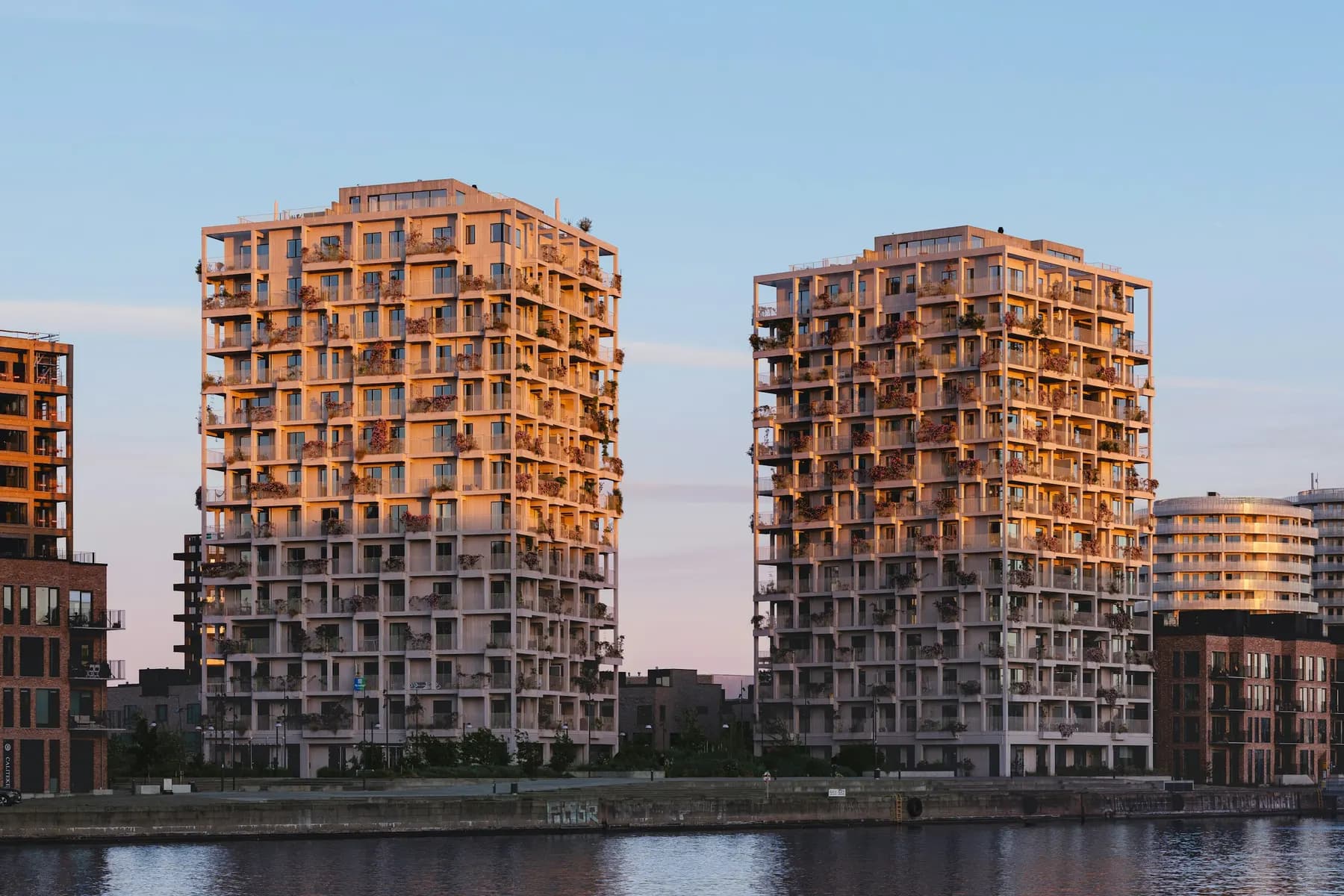 Deux tours résidentielles aux balcons végétalisés au bord de l'eau, éclairées par le soleil couchant.